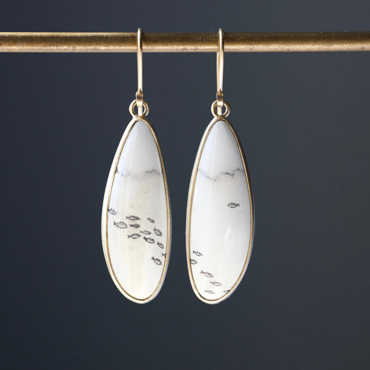 A pair of gold drop earrings with teardrop-shaped white stone inlays featuring small black fish motifs, hanging from a gold rod against a dark gray background.
