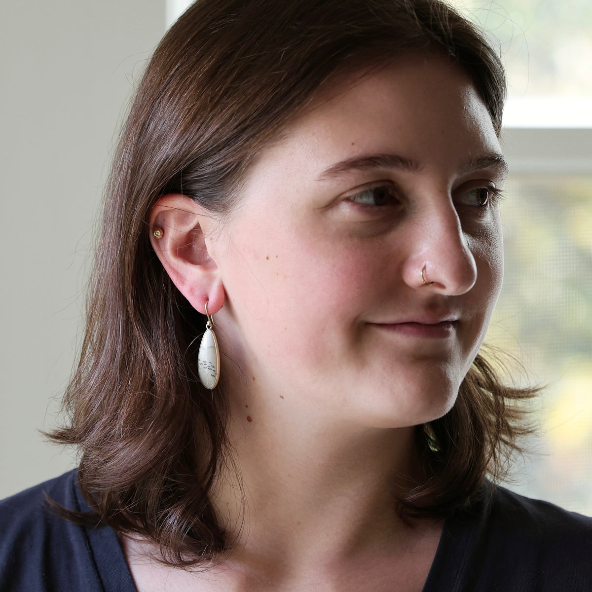 A woman with shoulder-length brown hair wearing a small white teardrop dangle earring, shown in a close-up portrait against a softly blurred indoor background.