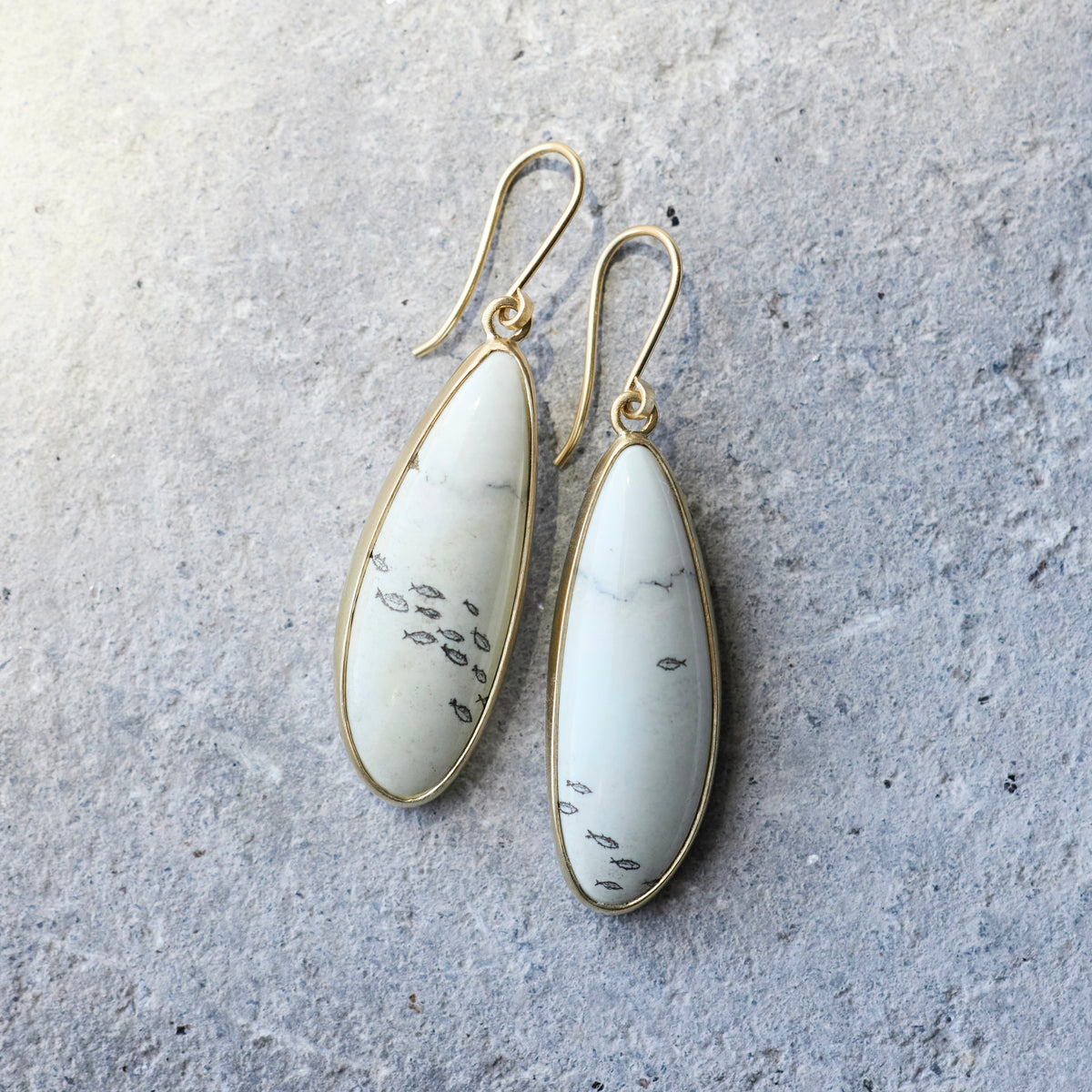 A pair of gold drop earrings with teardrop-shaped white stone inlays featuring black veining, displayed on a textured gray surface.