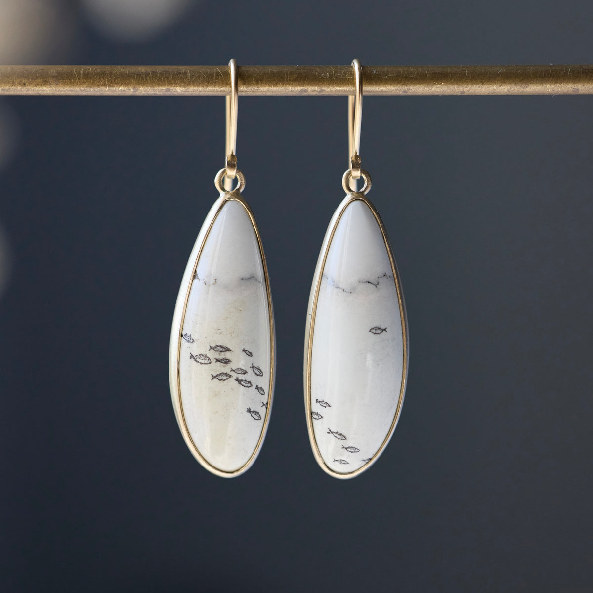 A pair of gold drop earrings with teardrop-shaped white stone pendants featuring black fish motifs, hanging from a gold rod against a dark gray background.
