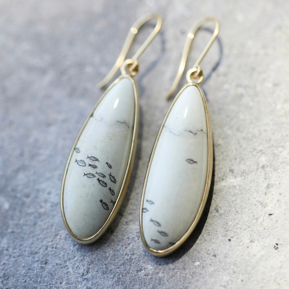 A pair of teardrop-shaped dangle earrings with white marbled stone inlays featuring small black fish motifs, set in gold-toned metal with matching gold fishhook ear wires, displayed on a textured gray surface.