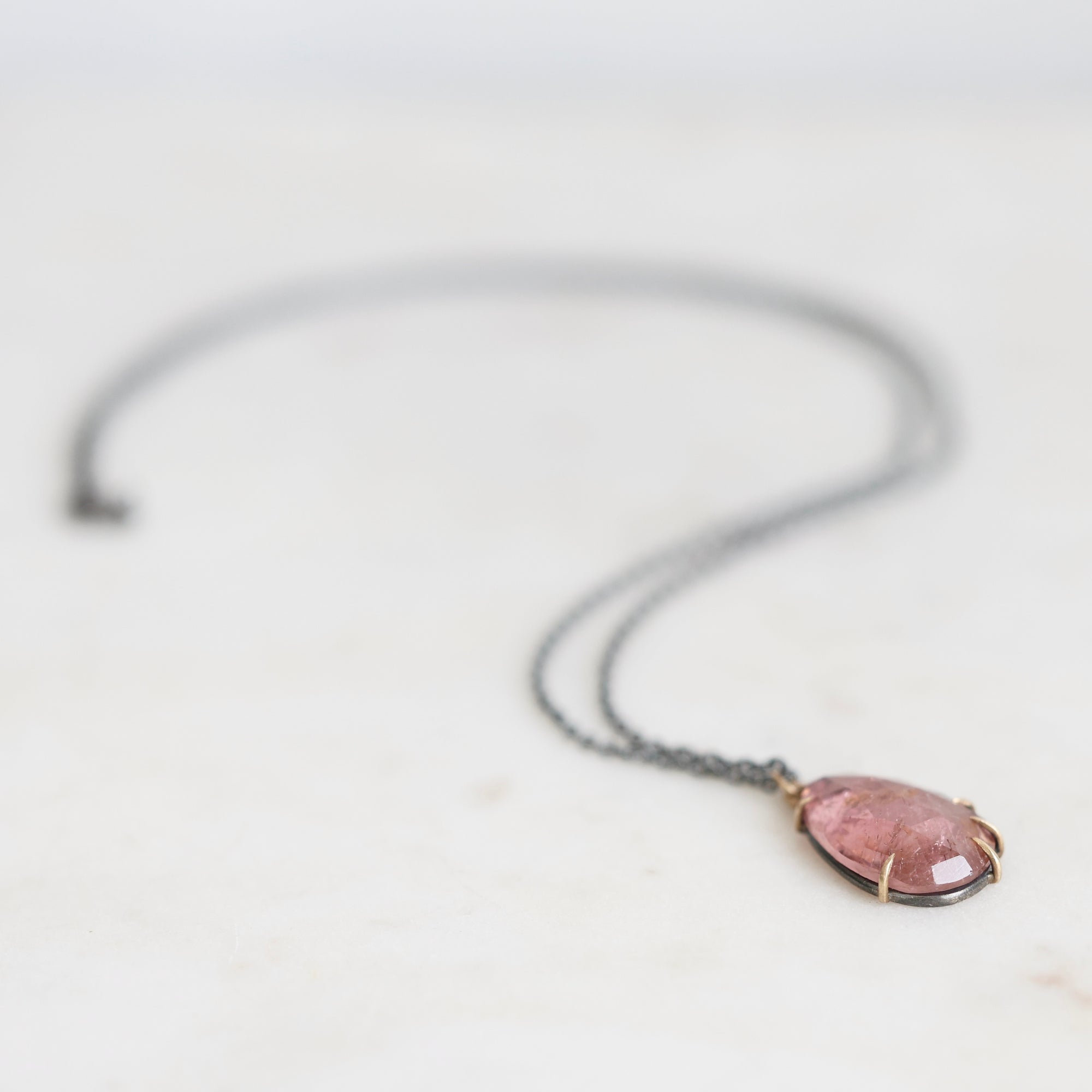 A delicate dark gray chain necklace with a small oval pendant featuring a pink stone in a gold-toned bezel setting, resting on a light off-white surface.