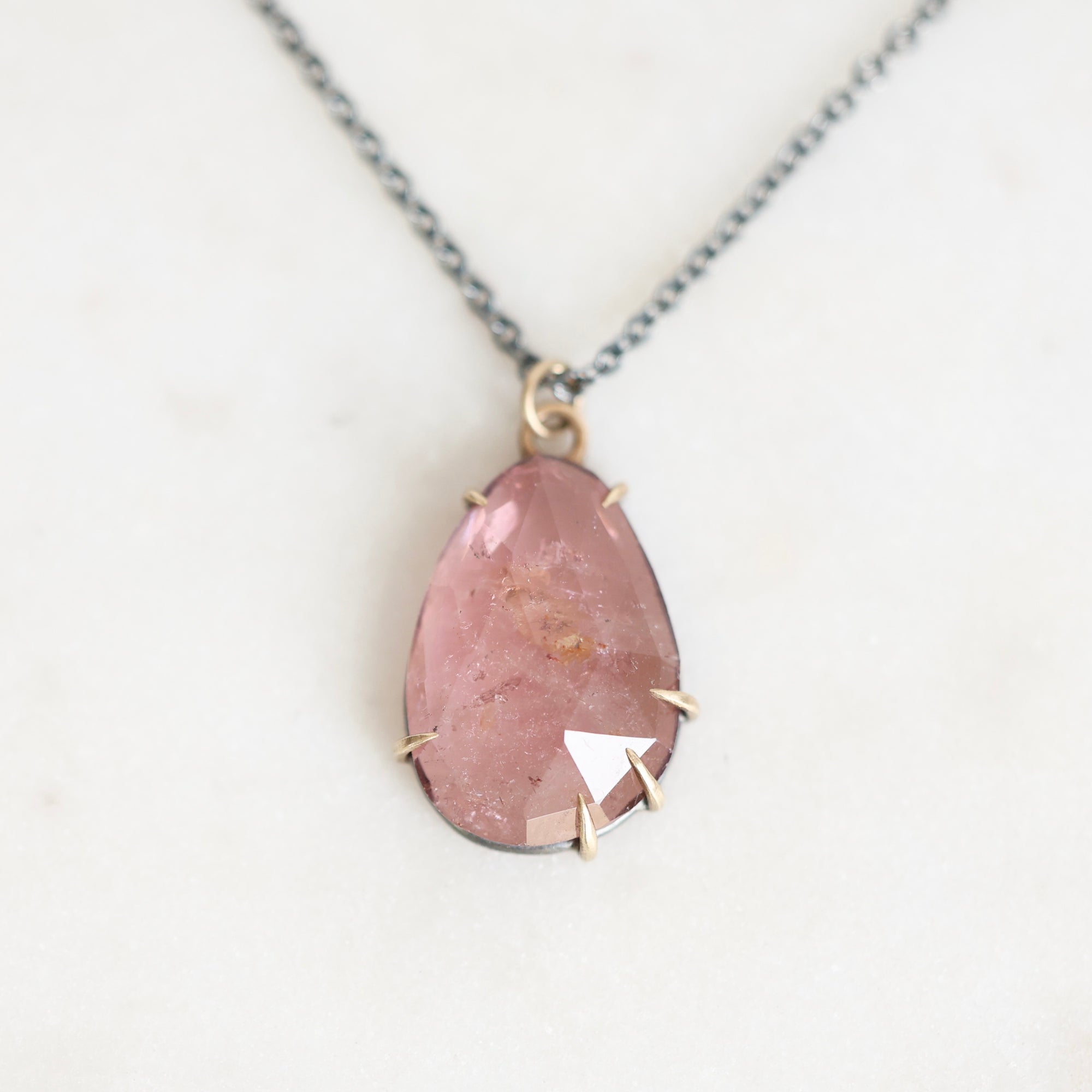A teardrop-shaped pink gemstone pendant necklace with a gold bezel setting and a silver-toned chain, displayed on a light off-white background.