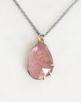A teardrop-shaped pink gemstone pendant necklace with a gold bezel setting and a silver-toned chain, displayed on a light off-white background.