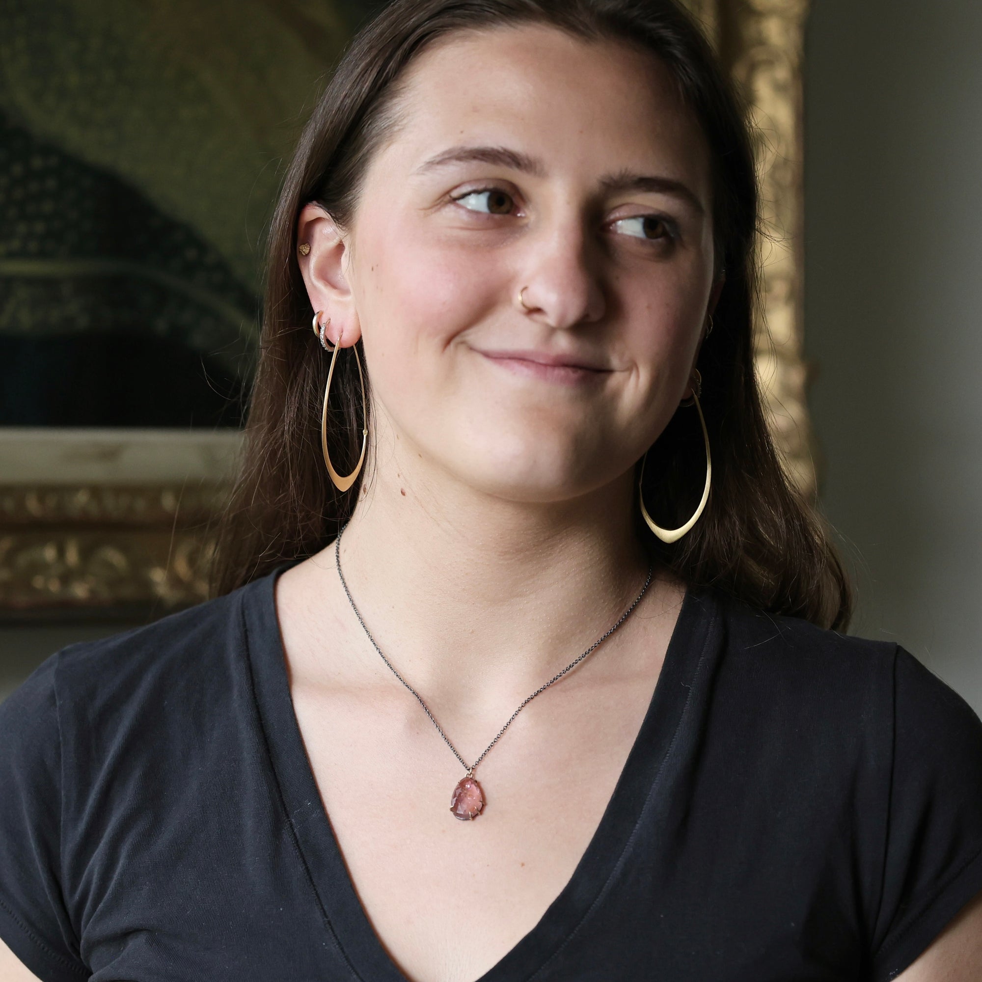 A woman with dark hair wearing large gold hoop earrings, a silver necklace with a pink teardrop pendant, and a black v-neck t-shirt, posing in front of a blurred ornate gold-framed picture.