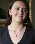 A woman with dark hair wearing large gold hoop earrings, a silver necklace with a pink teardrop pendant, and a black v-neck t-shirt, posing in front of a blurred ornate gold-framed picture.