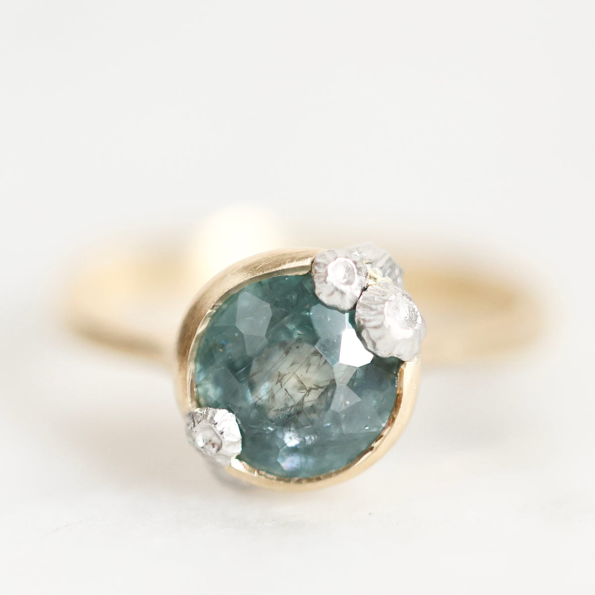 A gold ring featuring a large oval faceted blue-green gemstone with three small clear round-cut accent stones clustered at the top right, photographed on a clean white background.