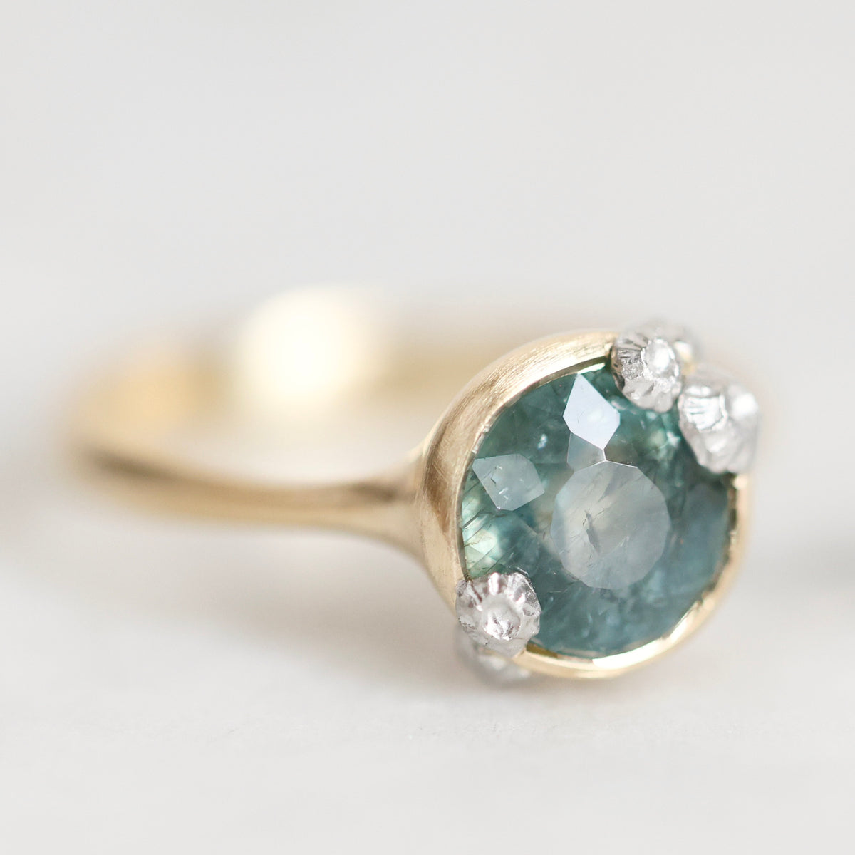A gold ring featuring a round faceted teal-blue gemstone in a prong setting, accented with three small clear diamonds, photographed on a soft light gray background.