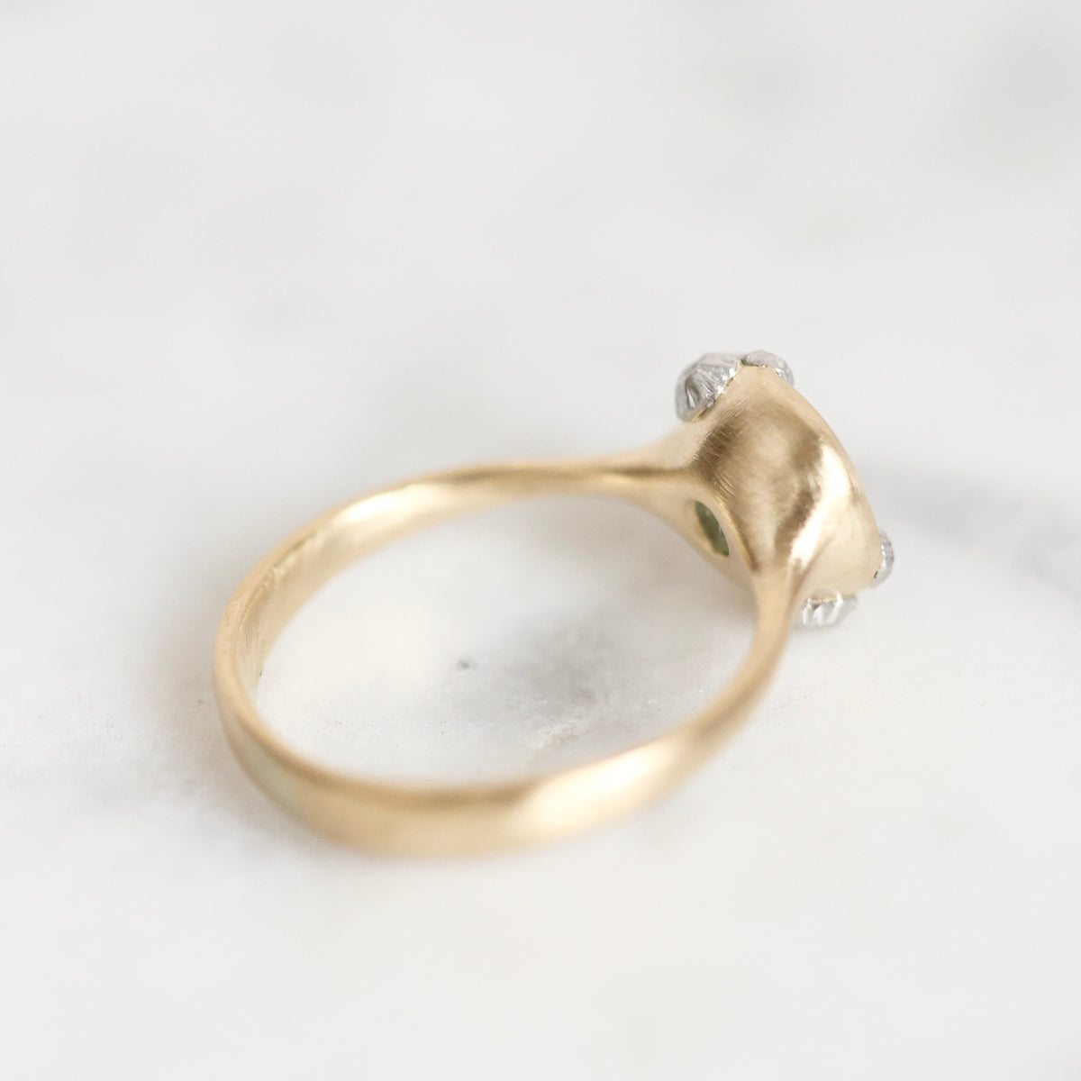 A gold ring with a sculptural, organic-shaped setting featuring small clear gemstones and a tiny green accent, photographed on a light marble surface.