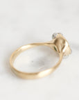 A gold ring with a sculptural, organic-shaped setting featuring small clear gemstones and a tiny green accent, photographed on a light marble surface.