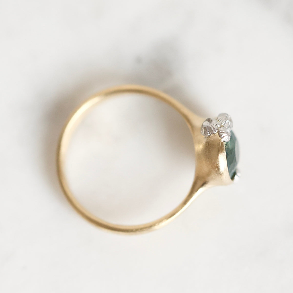 A gold ring with a small dark green gemstone and clear accent stones, photographed on a light marble surface.