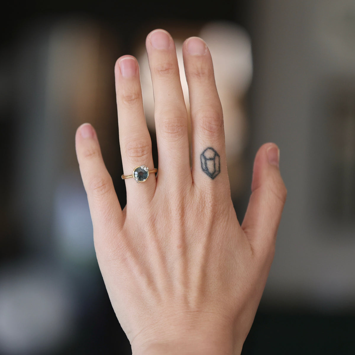 A close-up of a person's hand showing a gold ring with a dark green marquise-cut gemstone on the ring finger, alongside a small black geometric tattoo on the side of the hand.