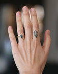 A close-up of a person's hand showing a gold ring with a dark green marquise-cut gemstone on the ring finger, alongside a small black geometric tattoo on the side of the hand.