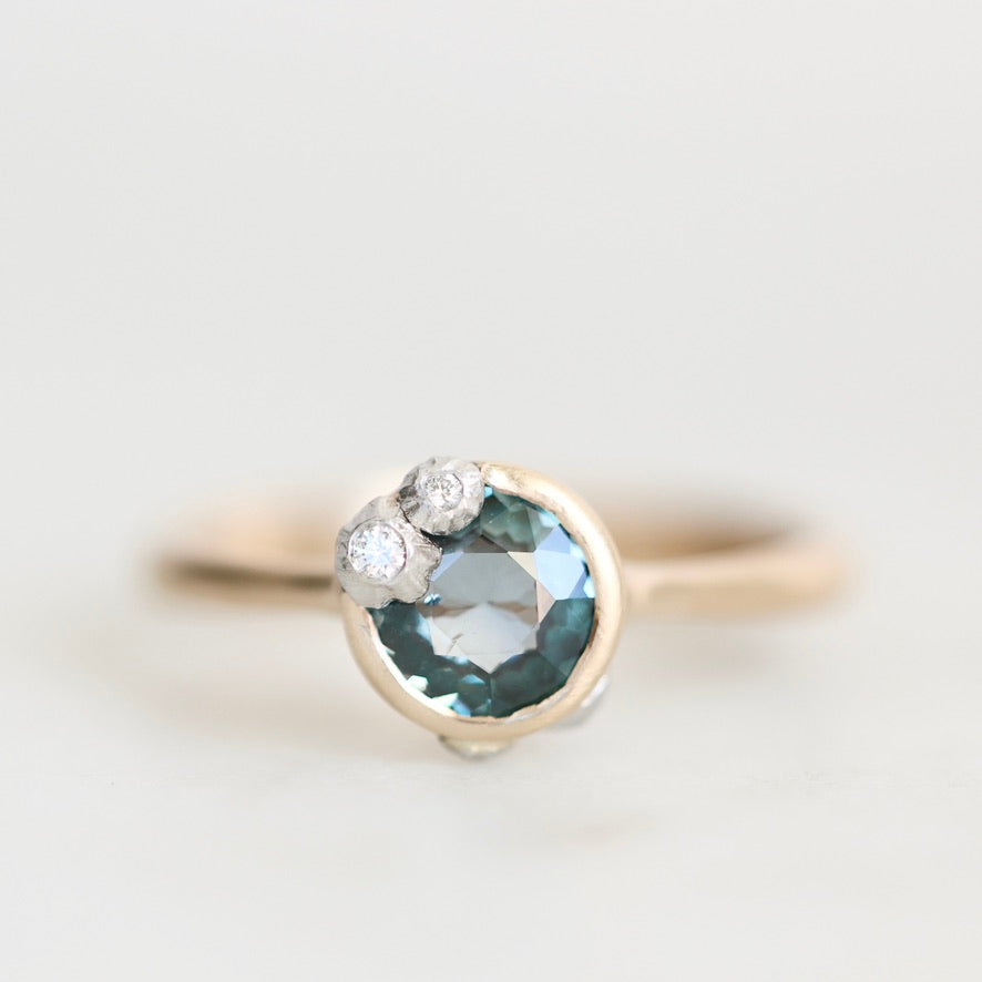 A close-up shot of a ring featuring a large oval-cut light blue gemstone in a gold bezel setting, with two small round-cut clear stones on the upper left. The ring is photographed on a clean, light gray background.