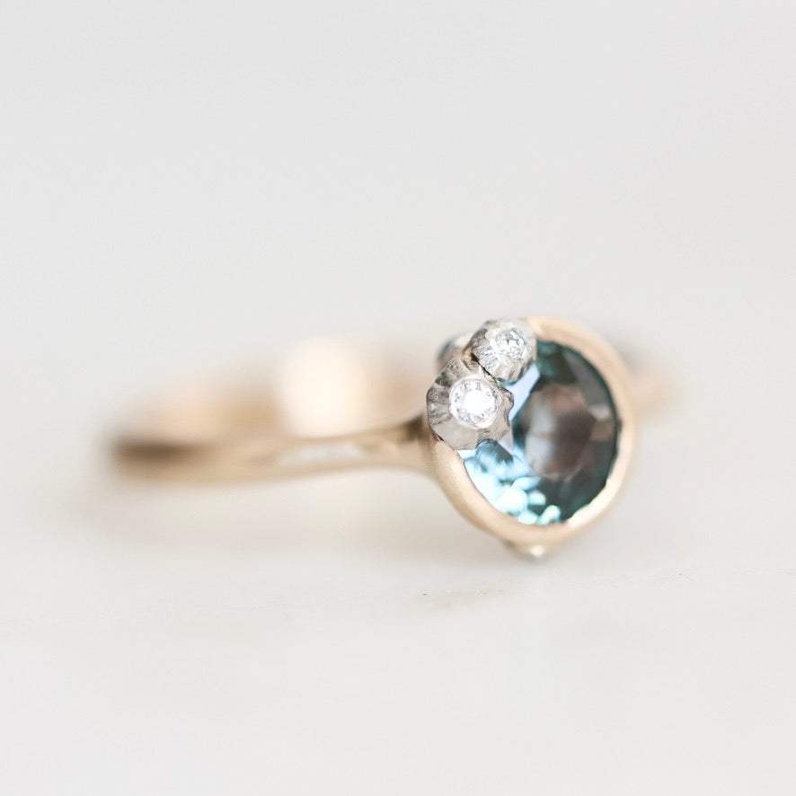 A close-up shot of a gold ring featuring a round light blue gemstone and three small clear accent stones, set against a soft, out-of-focus light background.