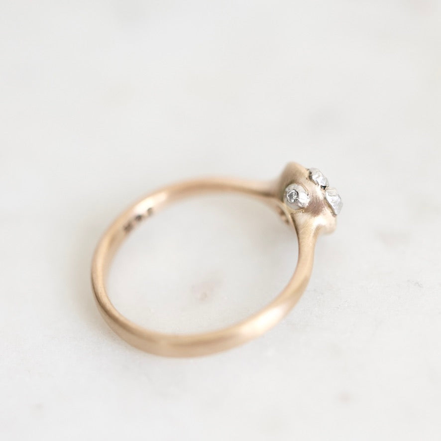 A close-up shot of a vintage-style ring with a gold band and a cluster of small clear gemstones on a light, off-white marble surface.