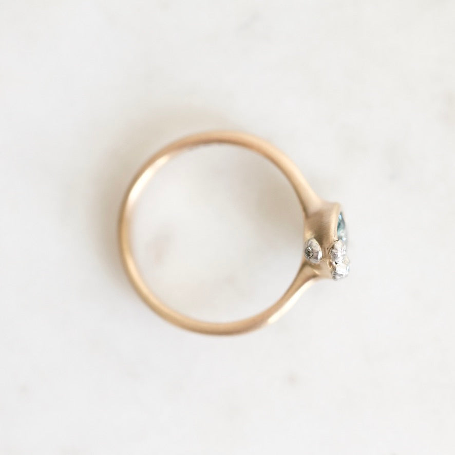 A gold ring with a small cluster of clear gemstones and a tiny light blue accent on the setting, photographed from above on a light off-white surface.