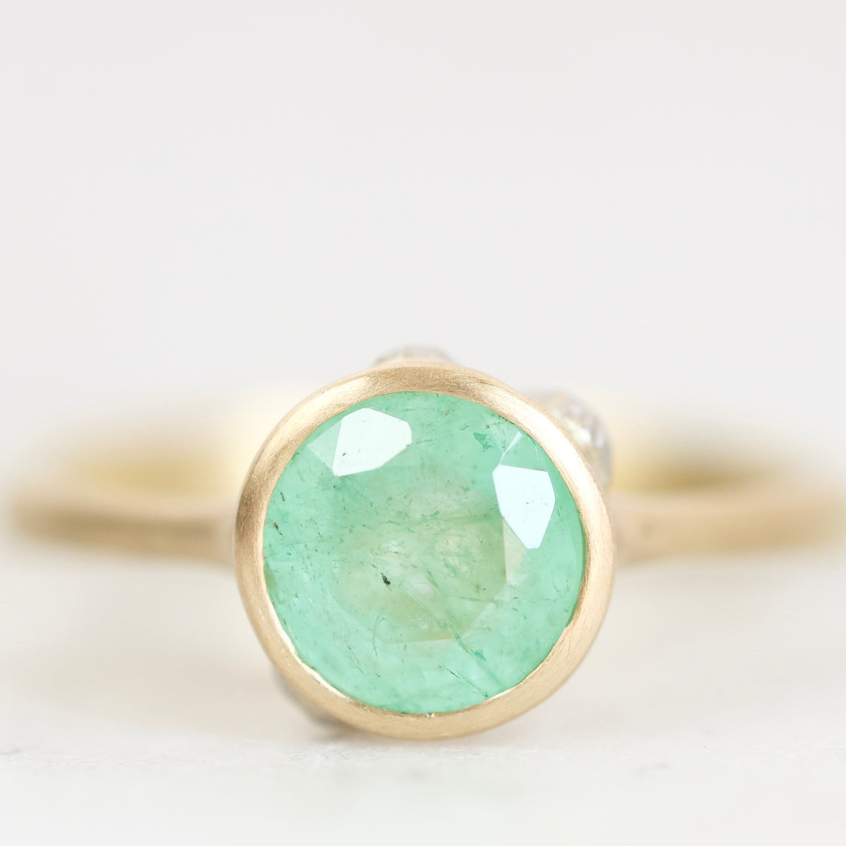 A gold ring featuring a round, faceted light green gemstone, photographed in a close-up studio shot against a soft, light gray background.