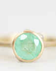 A gold ring featuring a round, faceted light green gemstone, photographed in a close-up studio shot against a soft, light gray background.