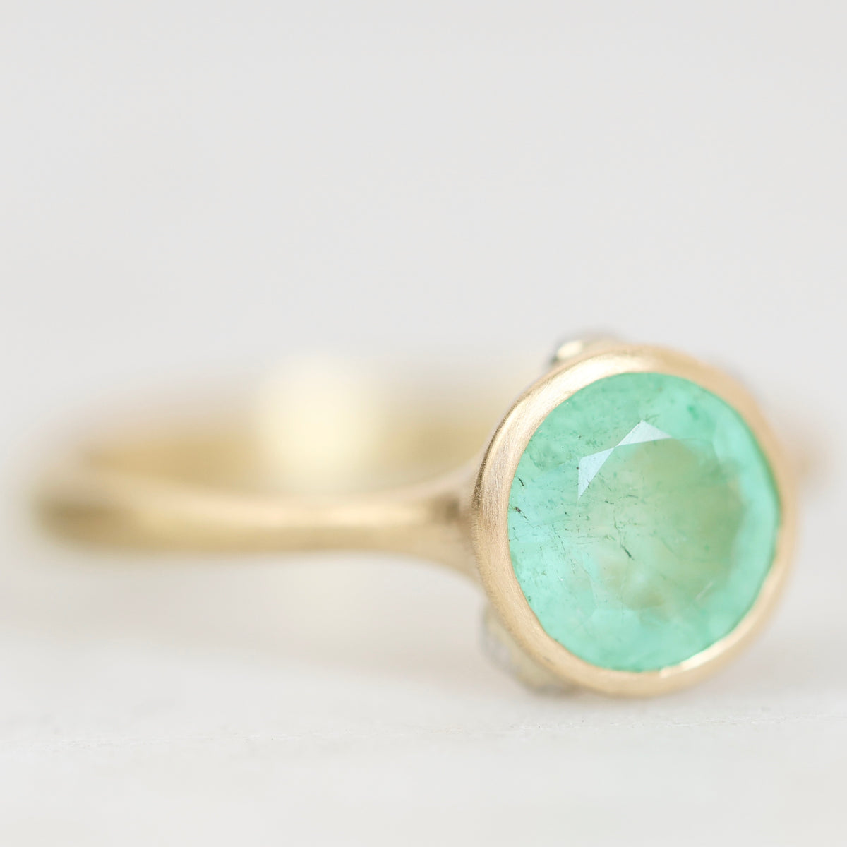A gold ring with a round faceted light green gemstone, shown in a close-up shot against a soft, light gray background.