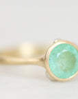 A gold ring with a round faceted light green gemstone, shown in a close-up shot against a soft, light gray background.