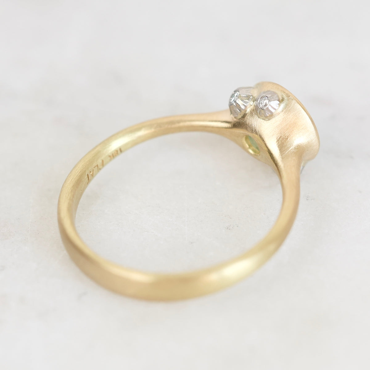 A close-up of a yellow gold ring with a textured, organic-shaped bezel setting holding two small clear gemstones, photographed on a light marble surface.