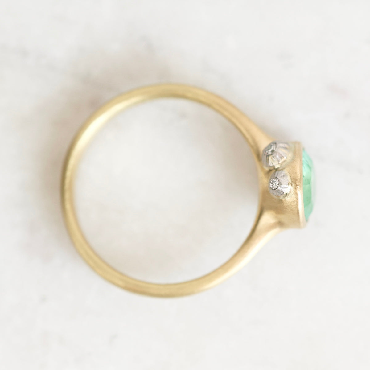 A gold ring with a bezel-set light green oval gemstone and two small clear stones, photographed on a light marble-like surface.