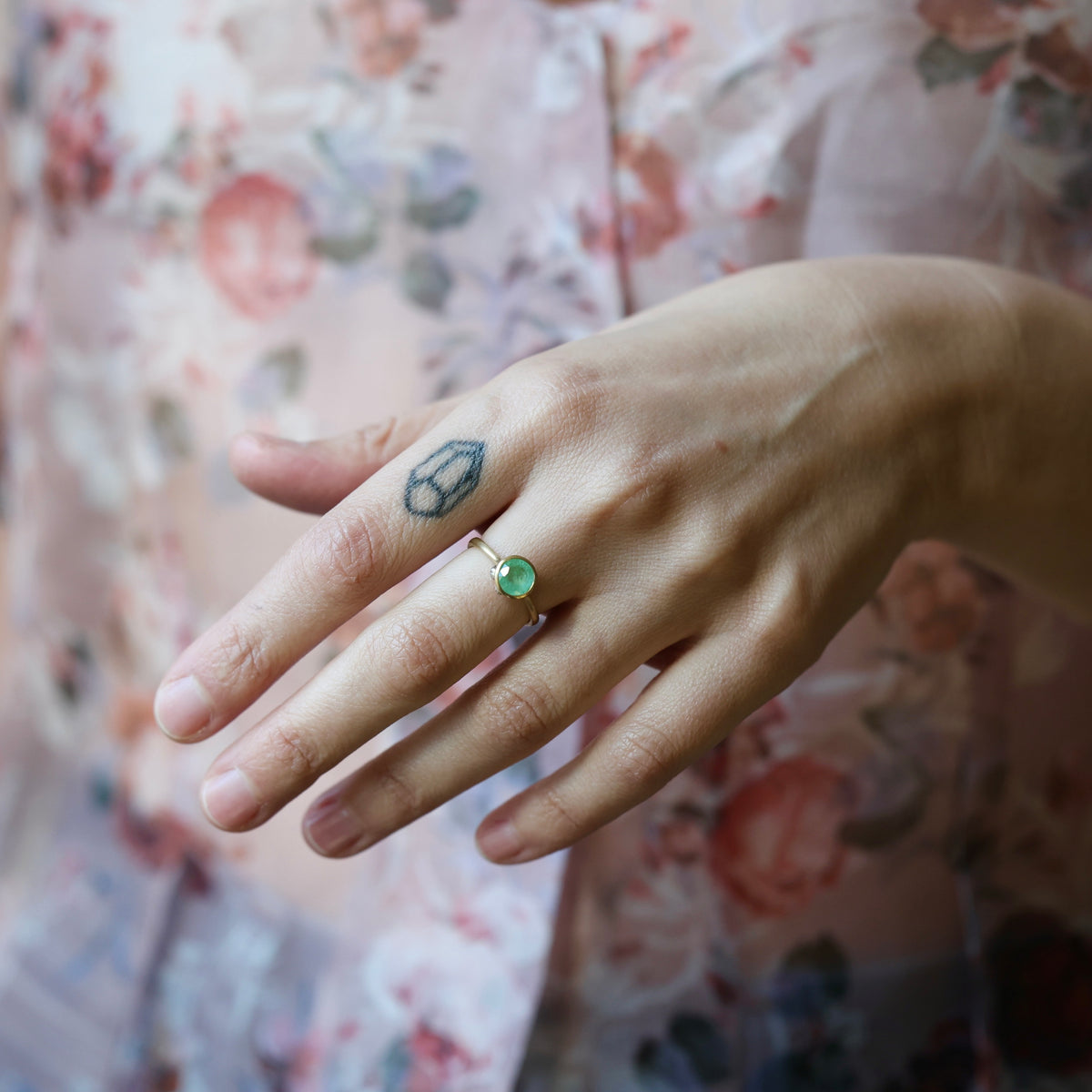 Hand wearing emerald bezel set ring against a floral blouse. 