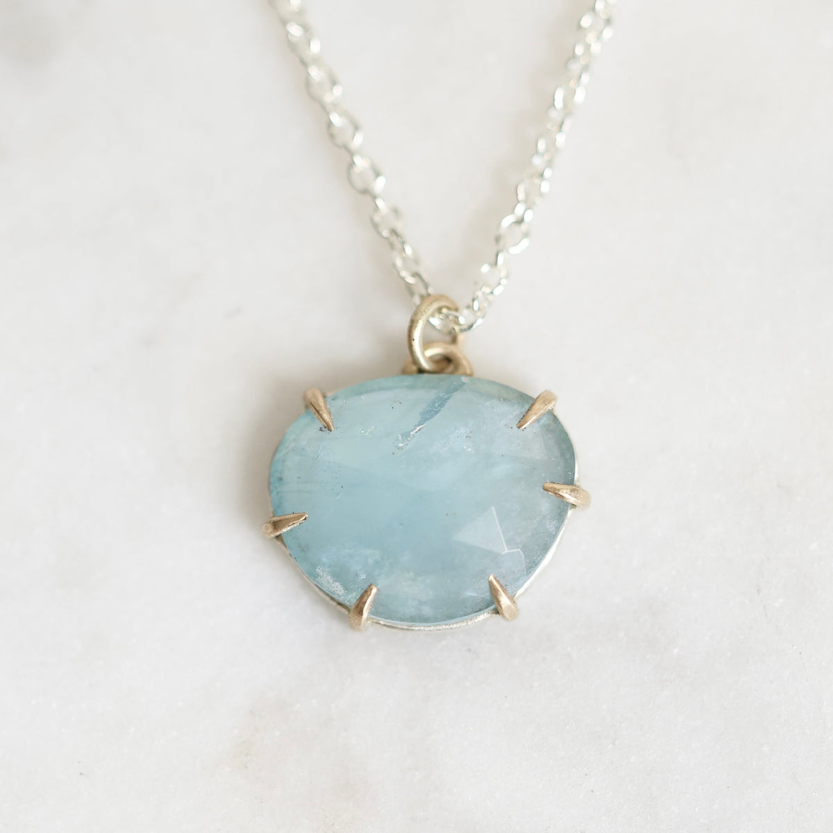 A light blue stone pendant necklace with a silver chain and a gold-toned prong setting, displayed on a light marble surface.