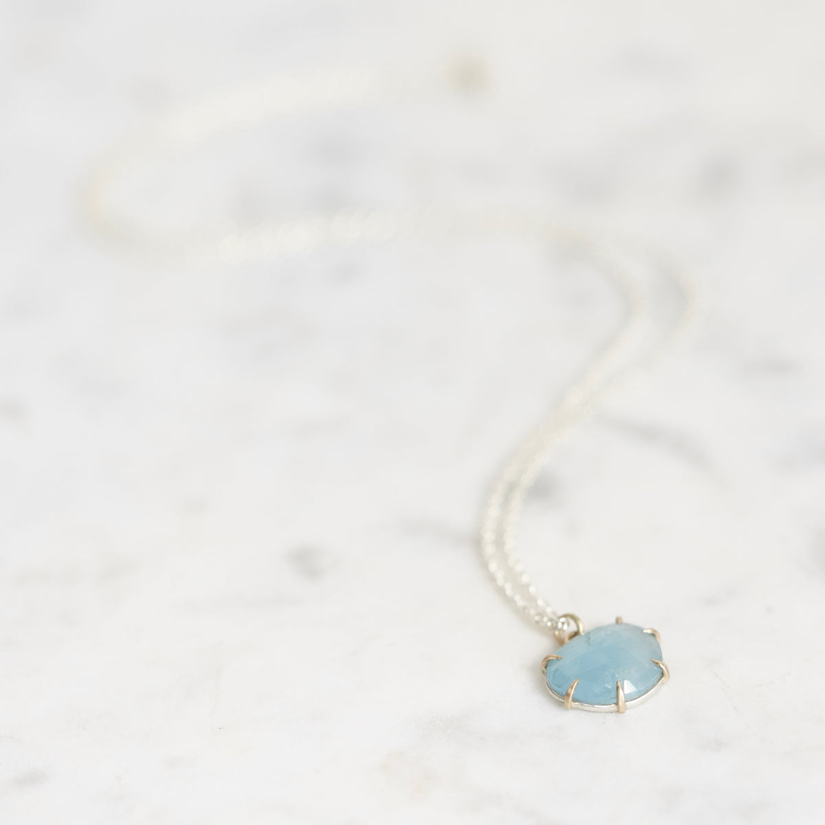A delicate silver chain necklace with a small oval pendant featuring a light blue stone, resting on a white marble surface.
