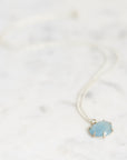 A delicate silver chain necklace with a small oval pendant featuring a light blue stone, resting on a white marble surface.