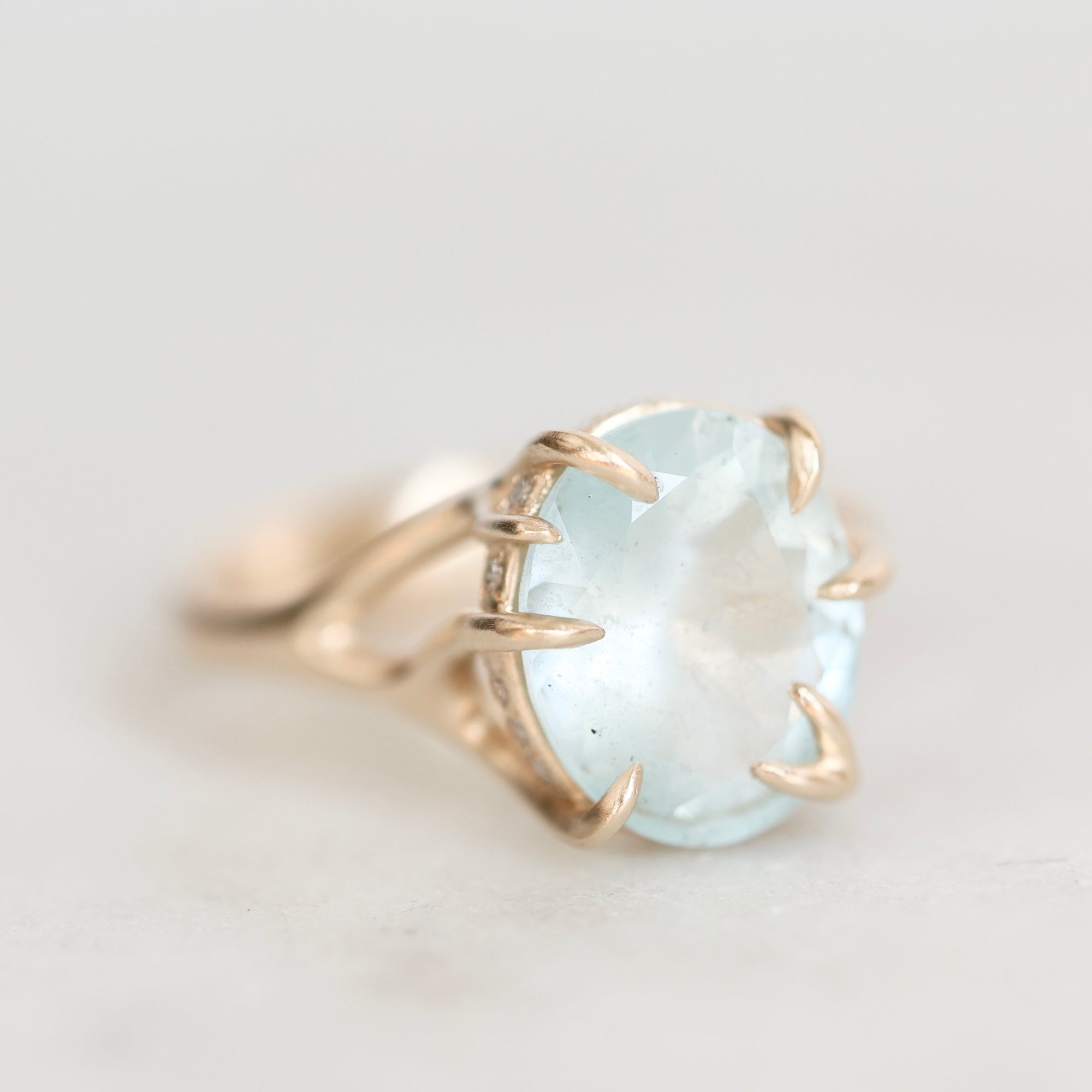 A gold ring featuring a large, oval-cut light blue gemstone in a claw setting, photographed on a soft, light gray background.