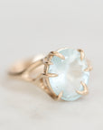 A gold ring featuring a large, oval-cut light blue gemstone in a claw setting, photographed on a soft, light gray background.