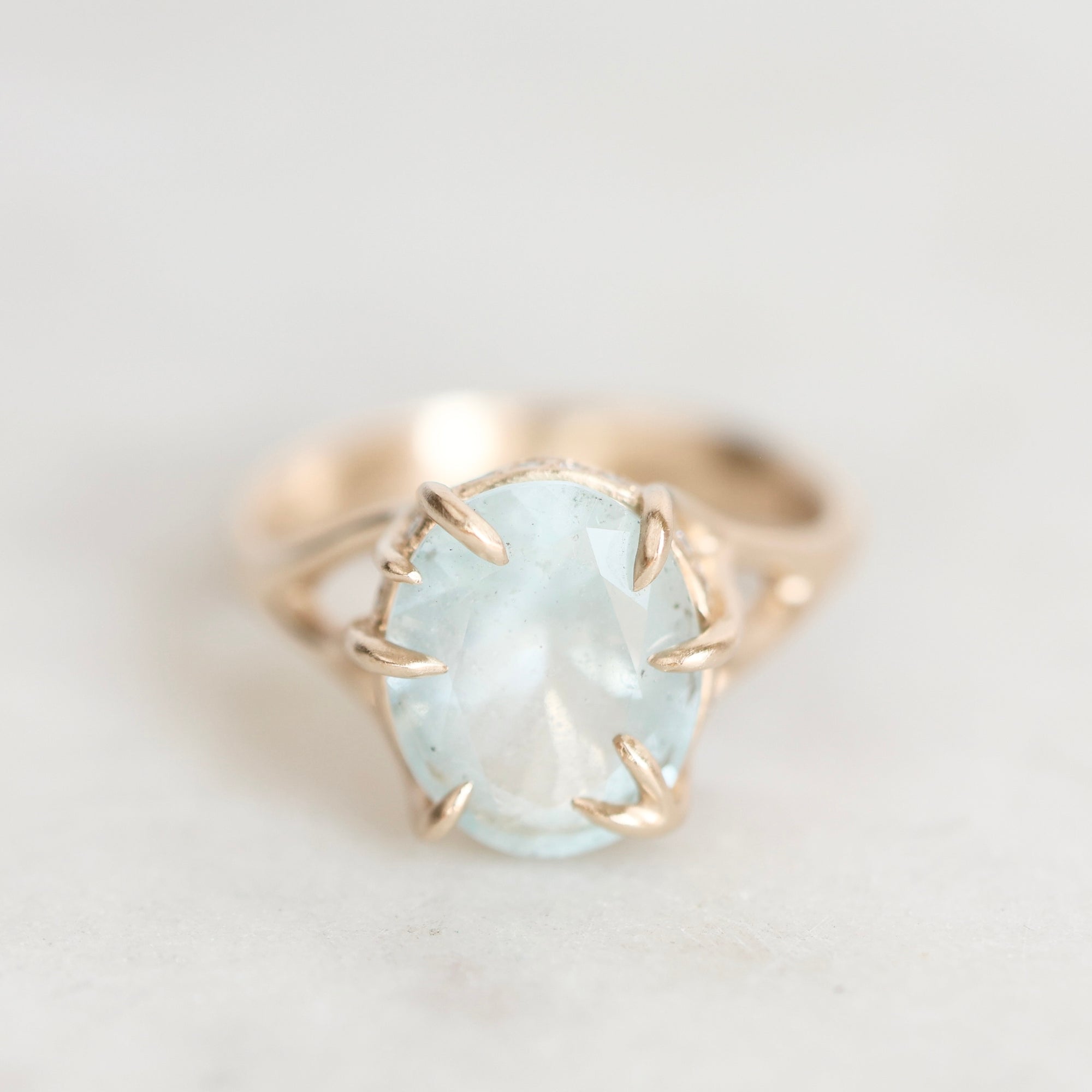 A gold ring featuring a large, oval-cut light blue gemstone in a prong setting, shown on a soft, light gray background.