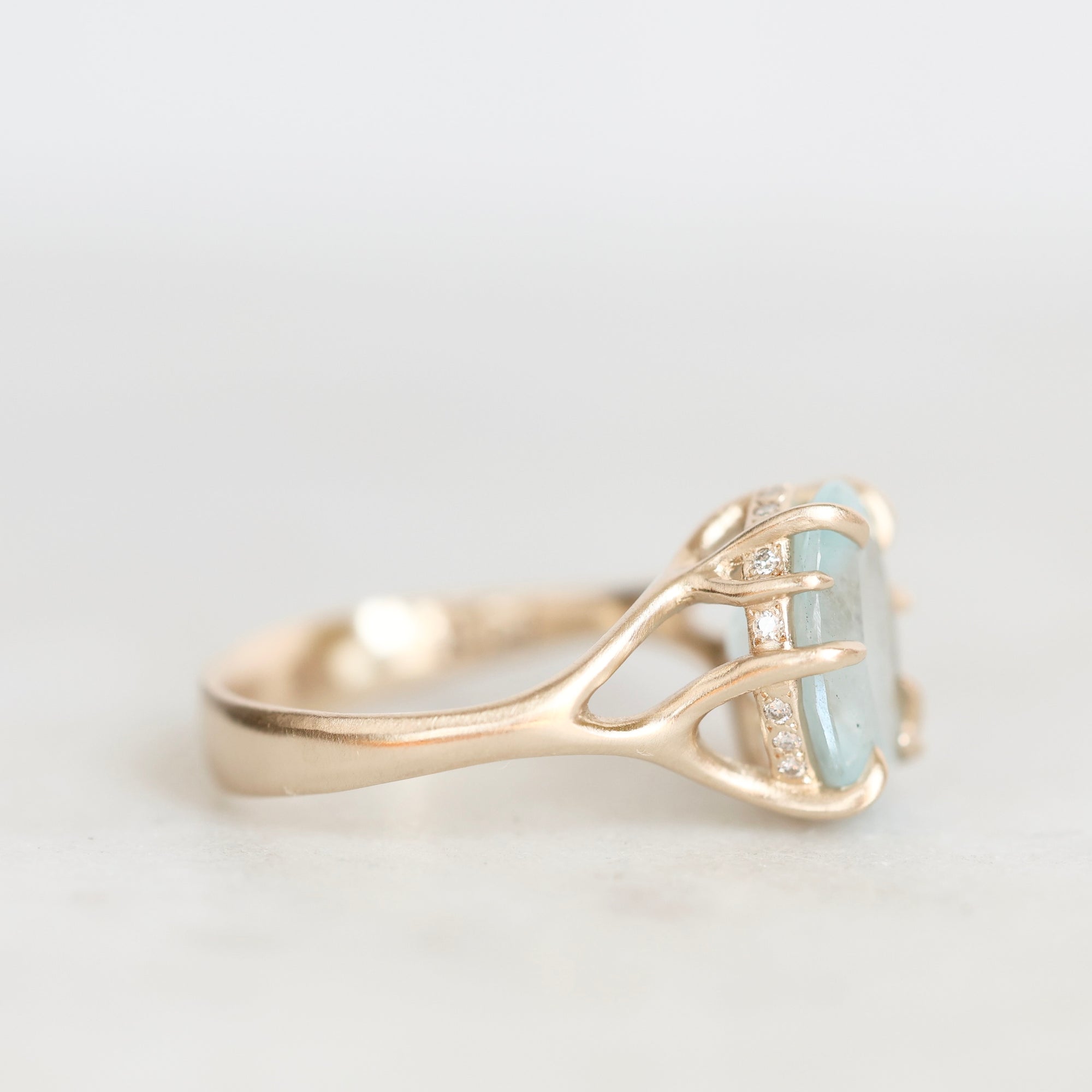 A gold ring with a unique antler-like setting holding a light blue rectangular gemstone, accented with small clear stones, shown on a light gray background.