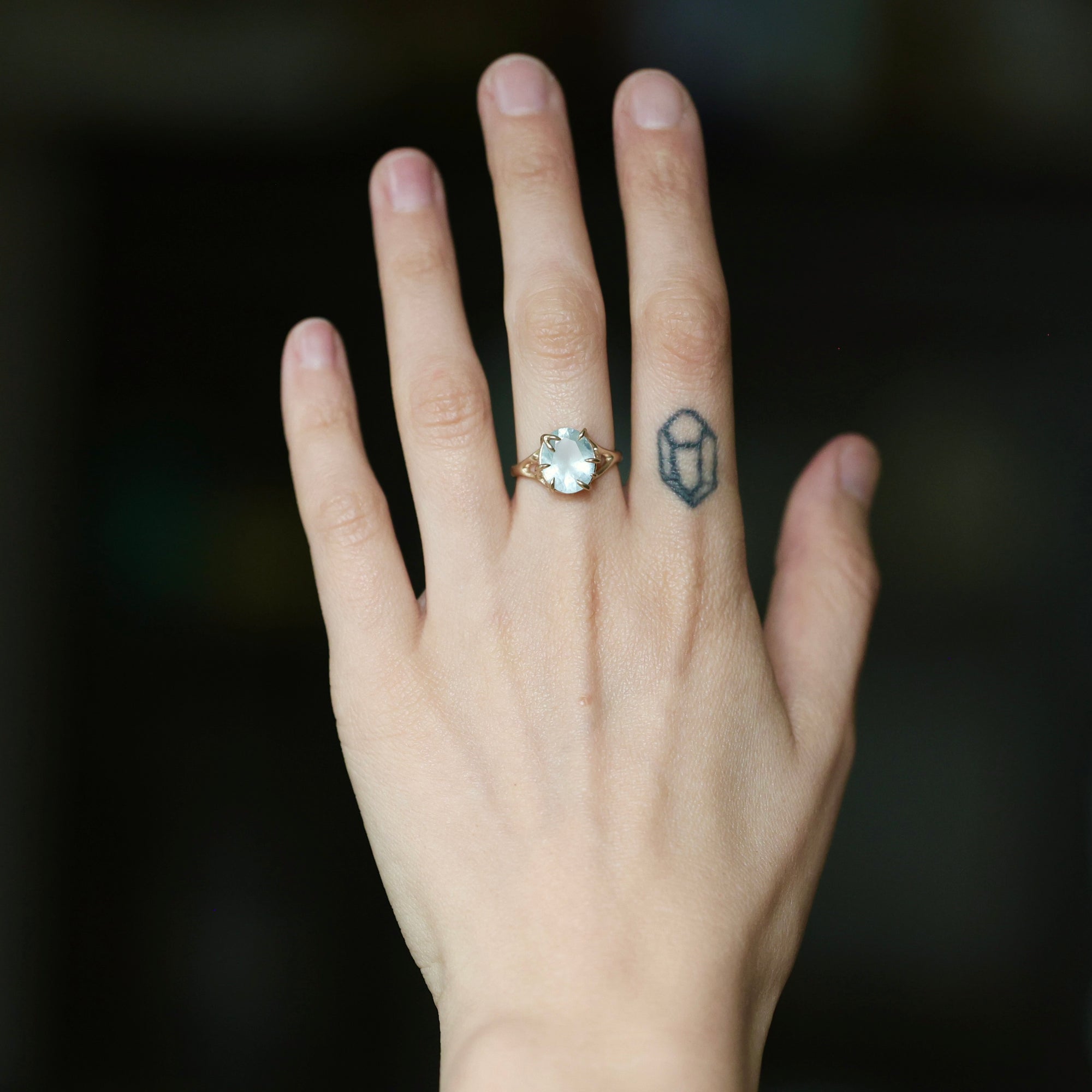 A close-up of a person's hand wearing a gold ring with a round white stone, with a small geometric tattoo on the middle finger, against a dark blurred background.
