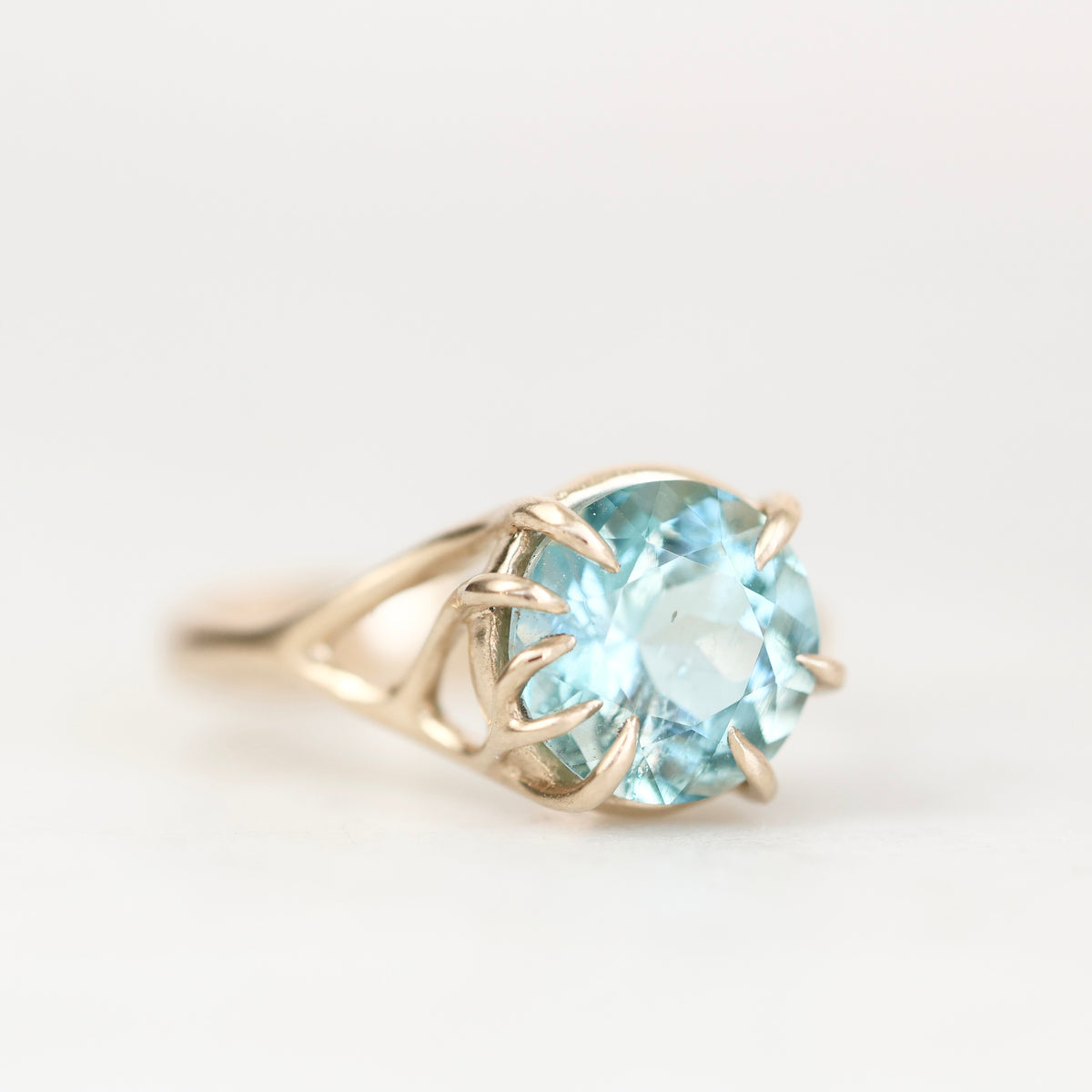 Blue gemstone ring with branchy gold prong setting against a plain background. 