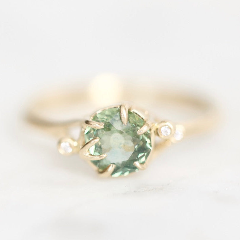 A yellow gold ring featuring a round green gemstone in a prong setting, accented by two small round clear stones on the shoulders, set against a soft white background.