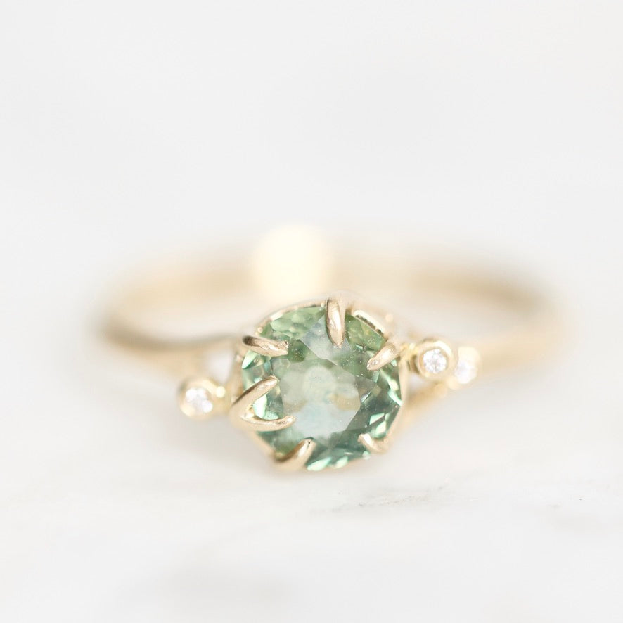 A yellow gold ring featuring a round green gemstone in a prong setting, accented by two small round clear stones on the shoulders, set against a soft white background.