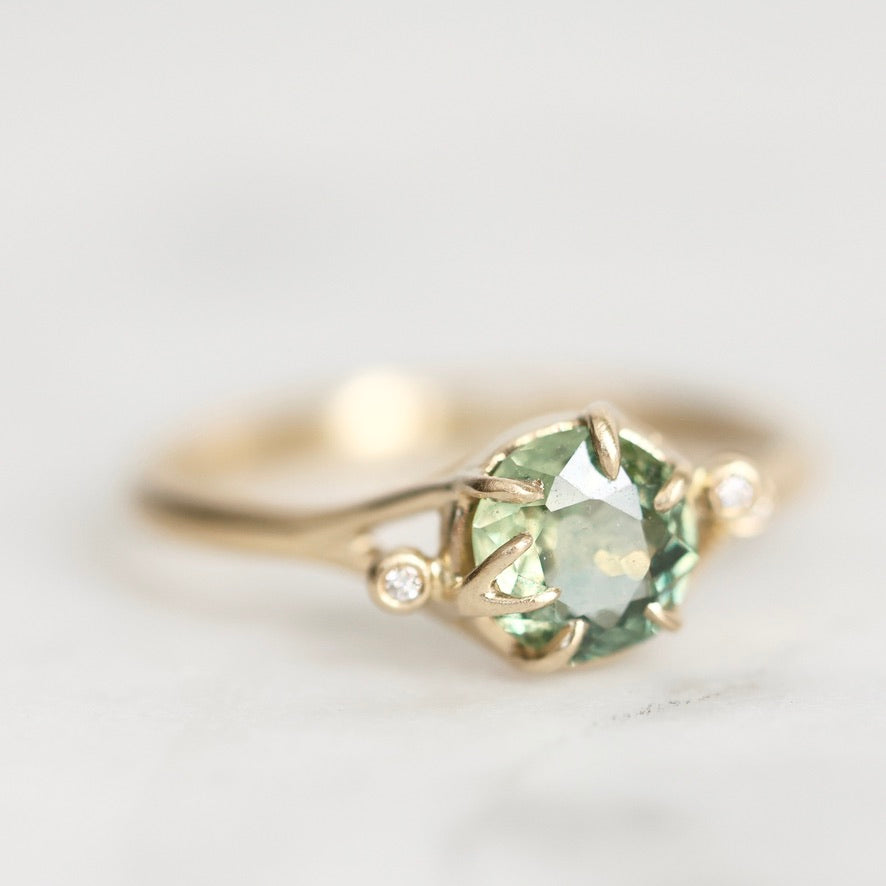 A gold ring featuring a round faceted light green gemstone in a prong setting, with two small round clear accent stones on the shoulders, photographed on a soft light gray background.