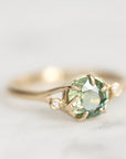 A gold ring featuring a round faceted light green gemstone in a prong setting, with two small round clear accent stones on the shoulders, photographed on a soft light gray background.
