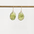 A pair of gold drop earrings with large, organic-shaped light green gemstones in prong settings, hanging from a gold wire stand against a plain white background.