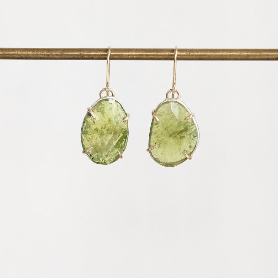 A pair of gold drop earrings with large, organic-shaped light green gemstones in prong settings, hanging from a gold wire stand against a plain white background.