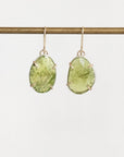 A pair of gold drop earrings with large, organic-shaped light green gemstones in prong settings, hanging from a gold wire stand against a plain white background.