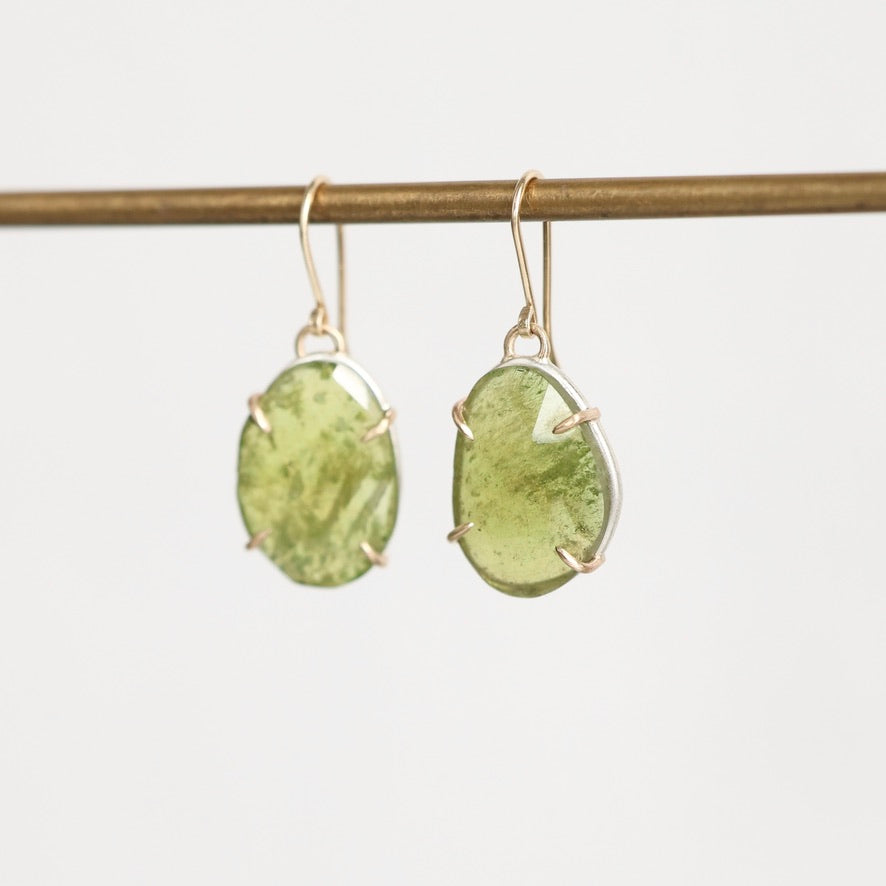 A pair of gold drop earrings with organic-shaped light green gemstones, hanging from a gold-colored horizontal bar against a plain white background.