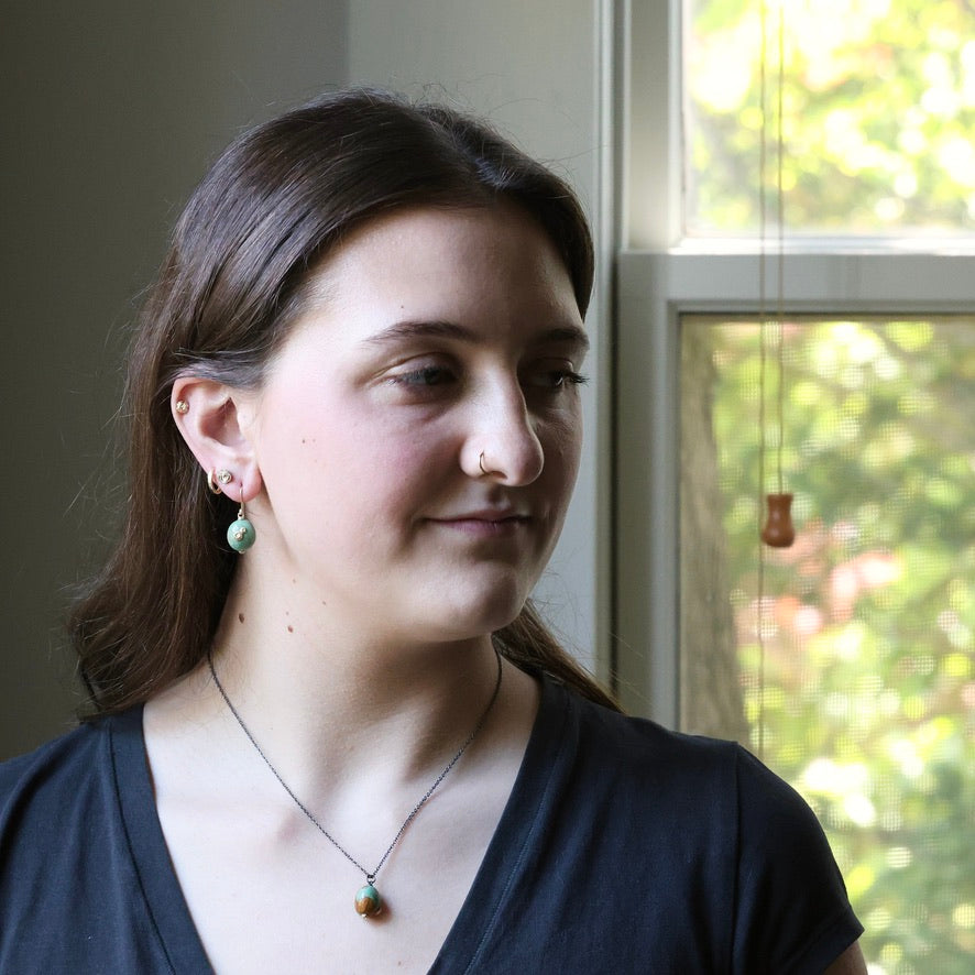 Woman wearing a necklace with a pendant, standing in front of a window with greenery outside.
