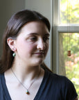 Woman wearing a necklace with a pendant, standing in front of a window with greenery outside.