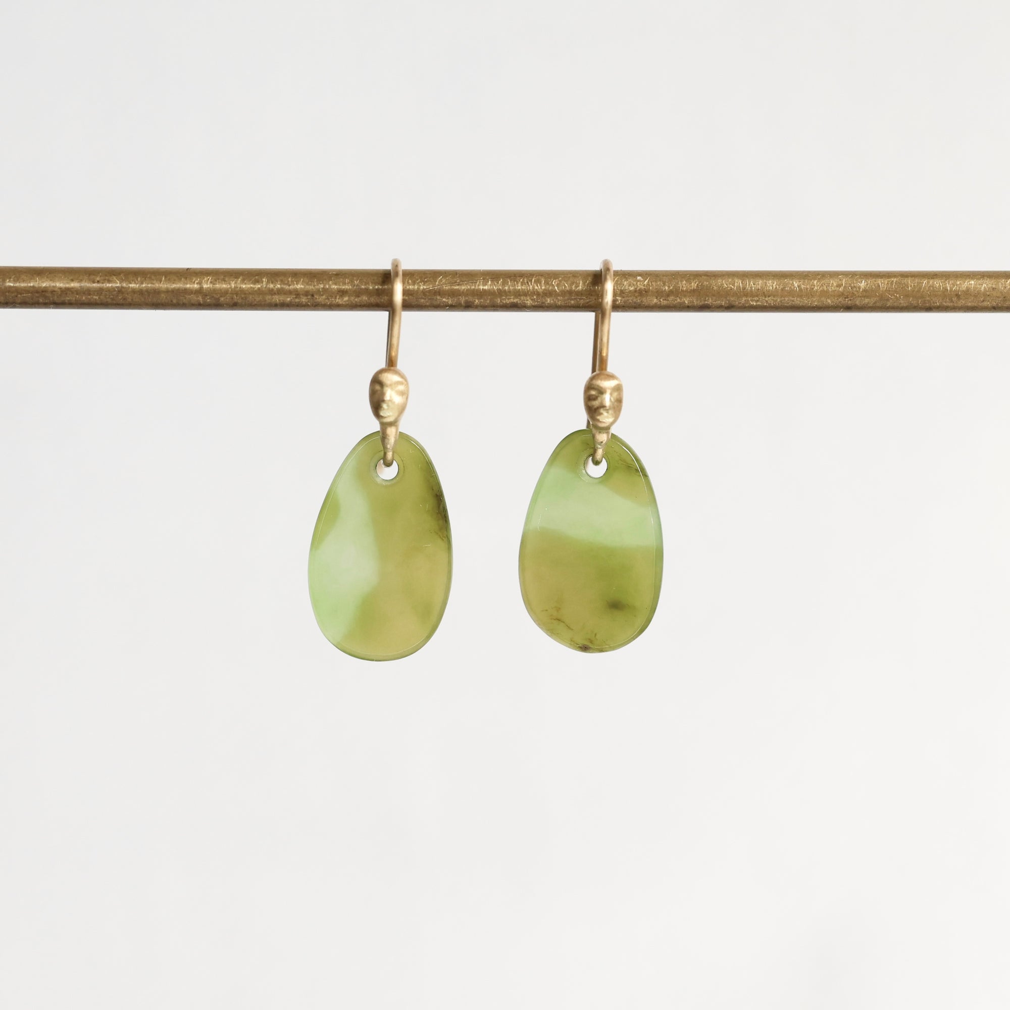 A pair of gold drop earrings with smooth, teardrop-shaped light green stone pendants, hanging from a horizontal gold rod against a plain white background.