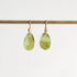 A pair of gold drop earrings with smooth, teardrop-shaped light green stone pendants, hanging from a horizontal gold rod against a plain white background.