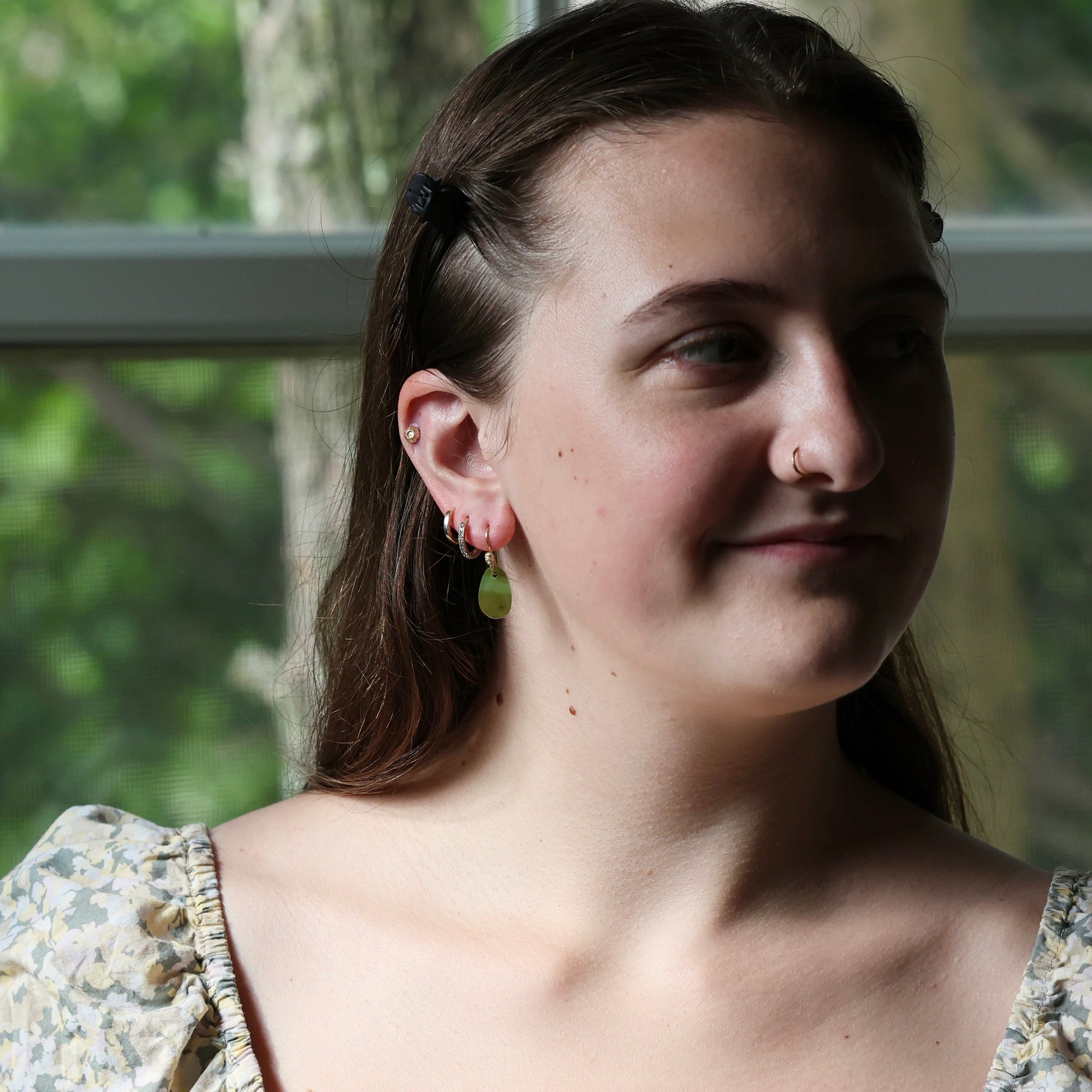 A close-up portrait of a woman wearing a small green teardrop dangle earring and a matching green hoop earring, photographed in natural light in front of a window.
